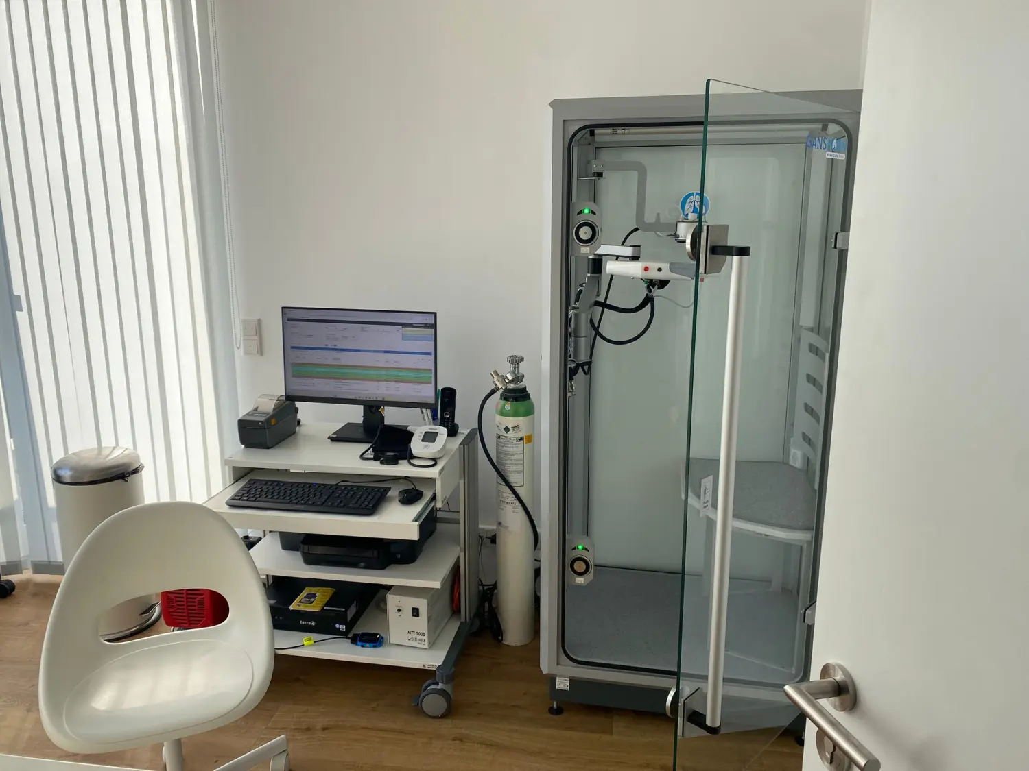 Facharztpraxis für Innere Medizin/Pneumologie – Diagnostik und Therapie - Untersuchungsraum mit HNO-Stuhl, Computer, Sauerstoffflasche und Rollstuhl